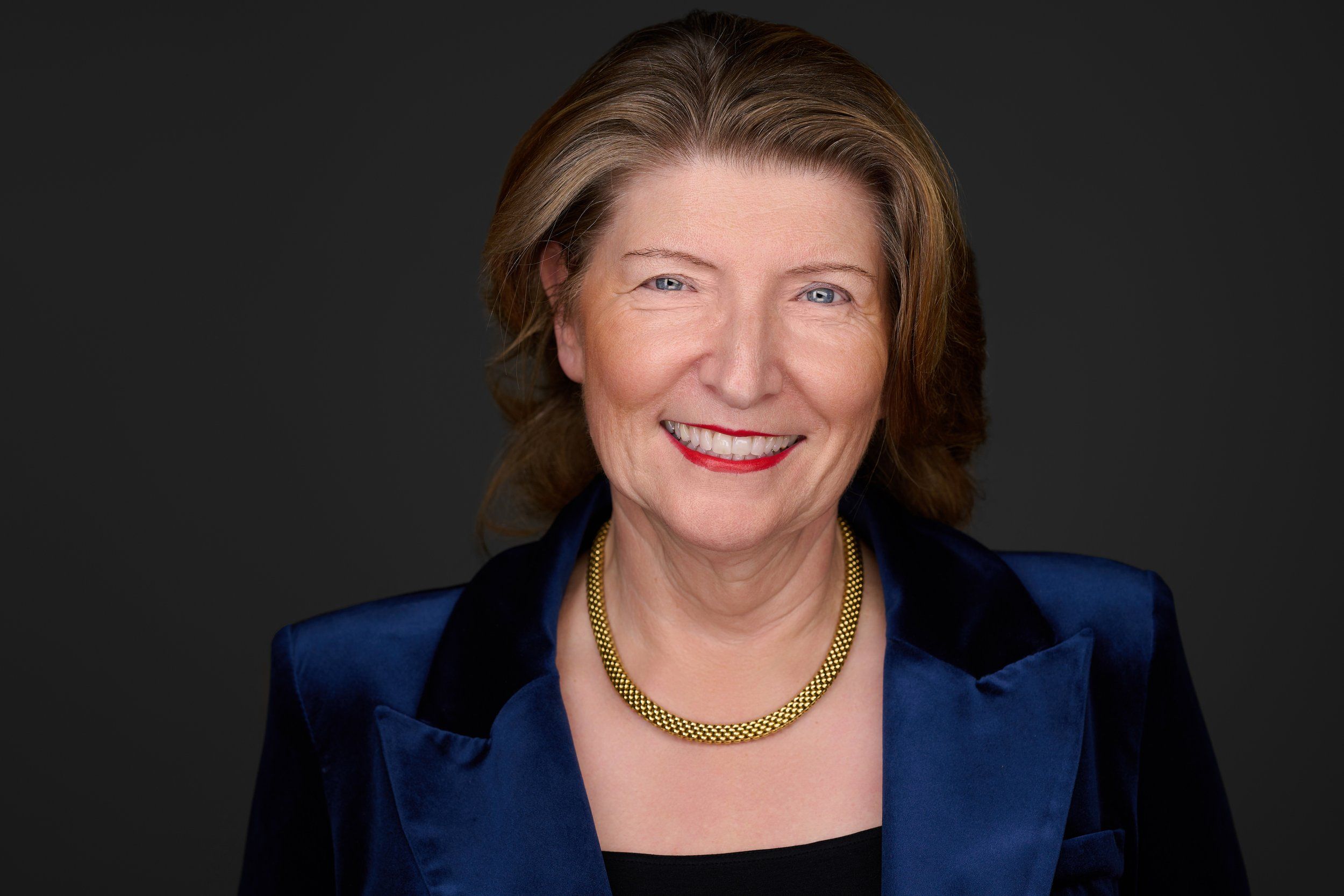 Headshot von Dr. Anna Martini: lächelnde Frau mit schulterlangen braunen Haaren, blauer Jacke und Goldkette vor dunklem Hintergrund