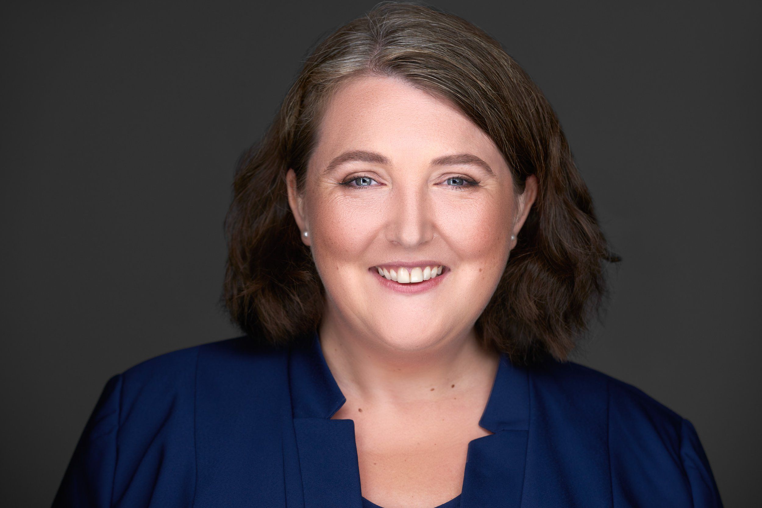 Businessportrait einer lächelnden Frau mit schulterlangen Haaren im blauen Blazer vor dunklem Hintergrund, fotografiert im Studio in Köln