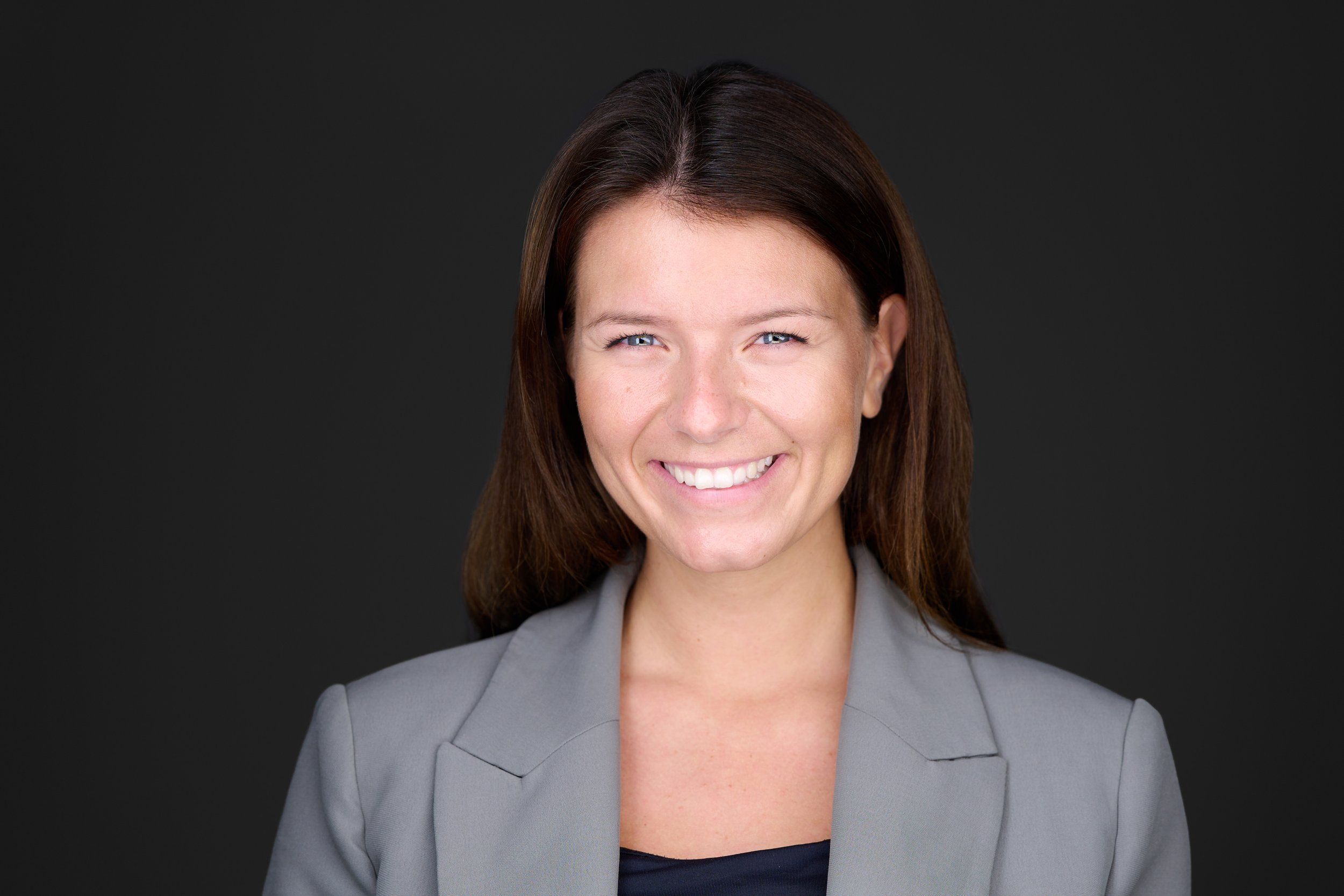 Businessportrait einer lächelnden Frau mit langen Haaren im grauen Blazer vor dunklem Hintergrund, fotografiert im Studio in Köln