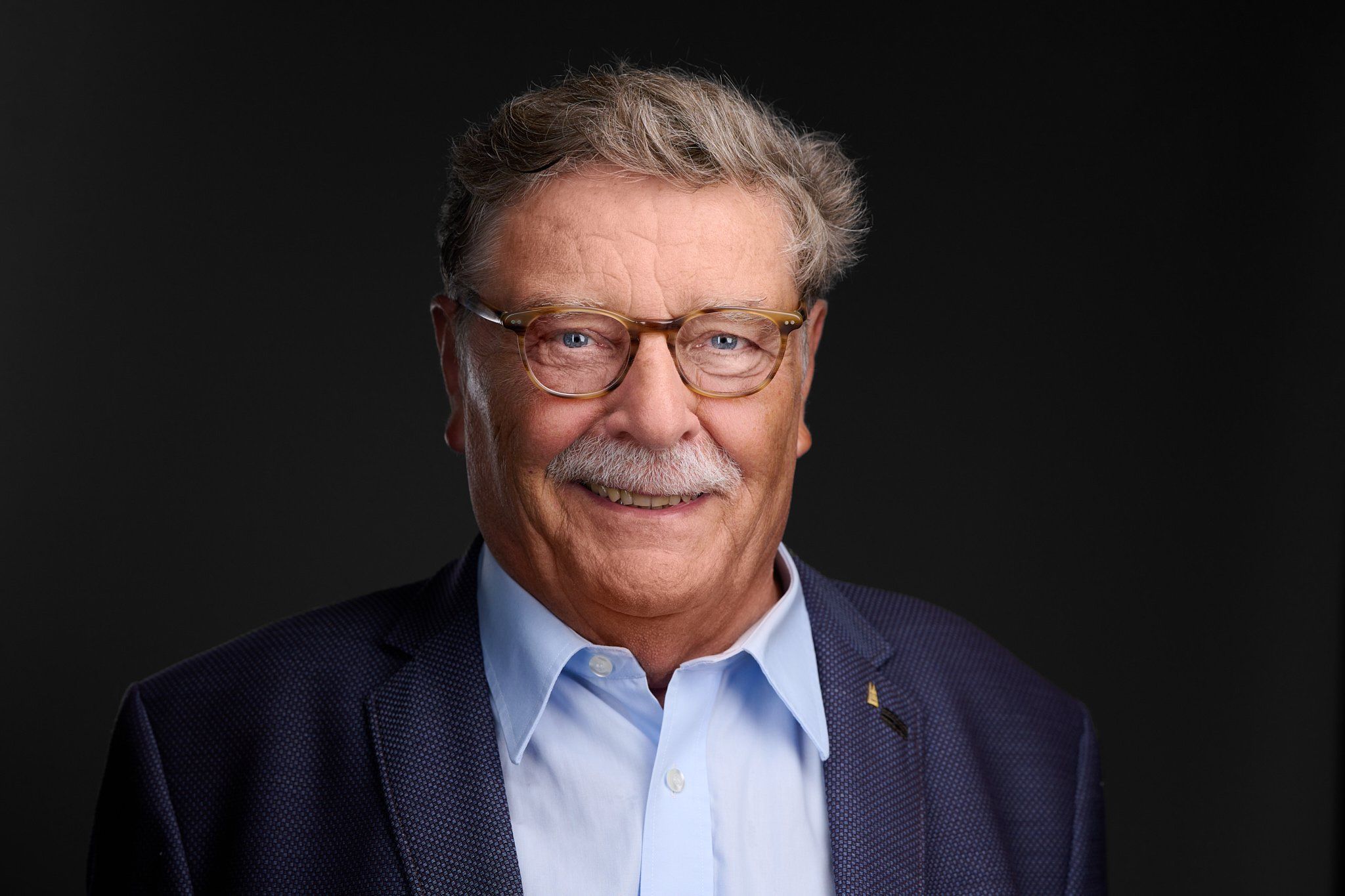 Headshot von Fritz Schramma: älterer Mann mit grauem Haar, Brille und blauem Sakko vor dunklem Hintergrund