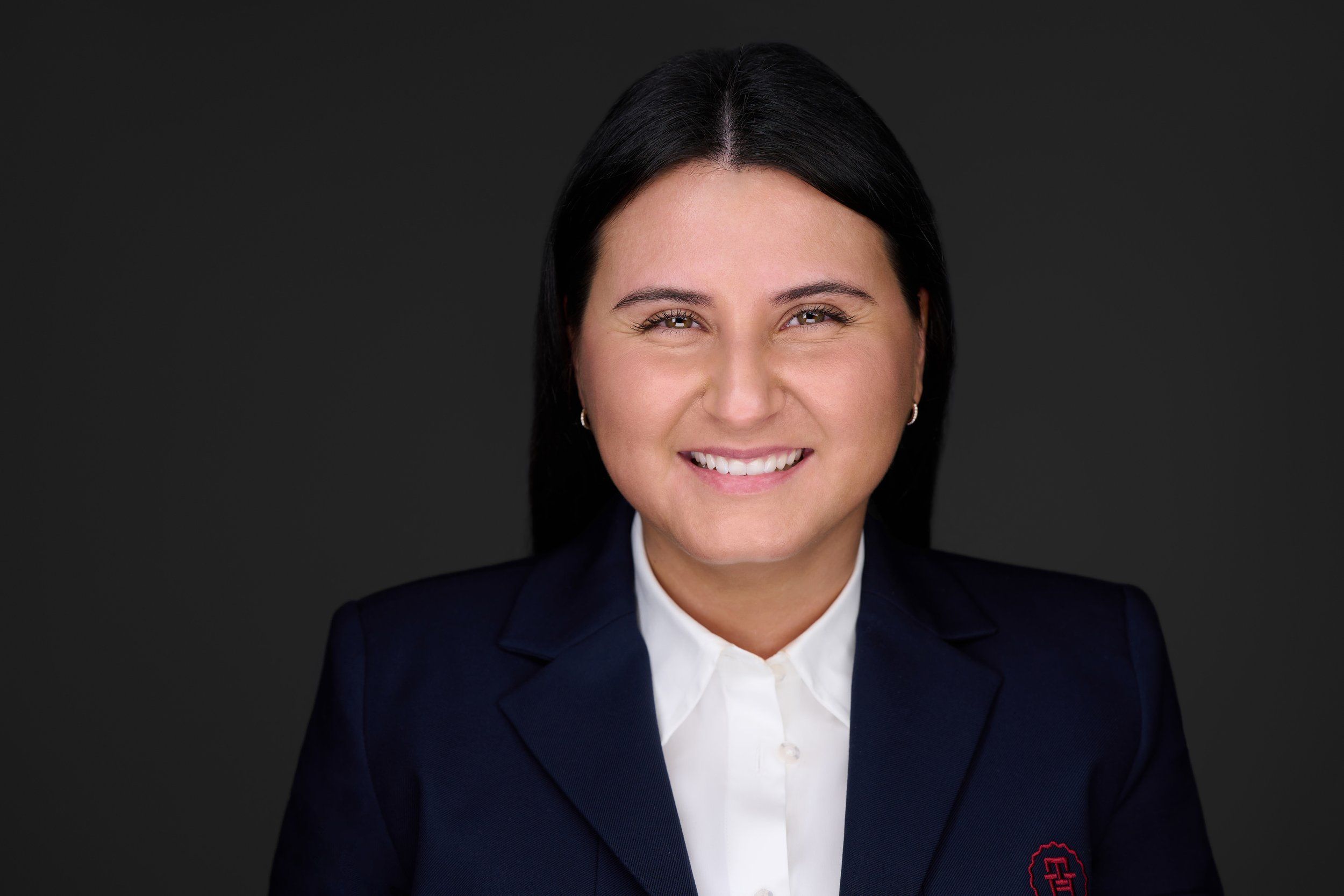 Businessportrait einer lächelnden jungen Frau mit langen schwarzen Haaren im dunkelblauen Blazer vor dunklem Hintergrund, fotografiert im Studio in Köln