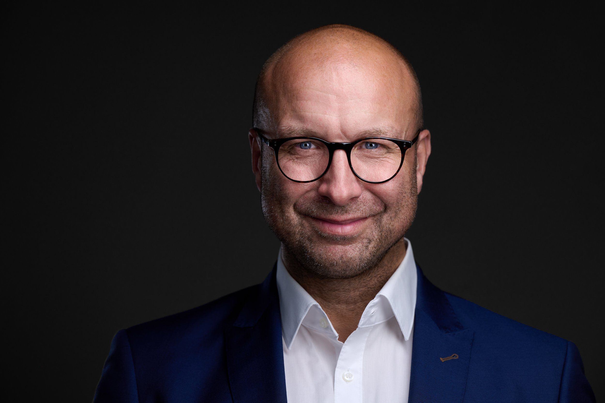 Businessportrait eines lächelnden Mannes mit Brille im dunkelblauen Anzug vor dunklem Hintergrund, fotografiert im Studio in Köln