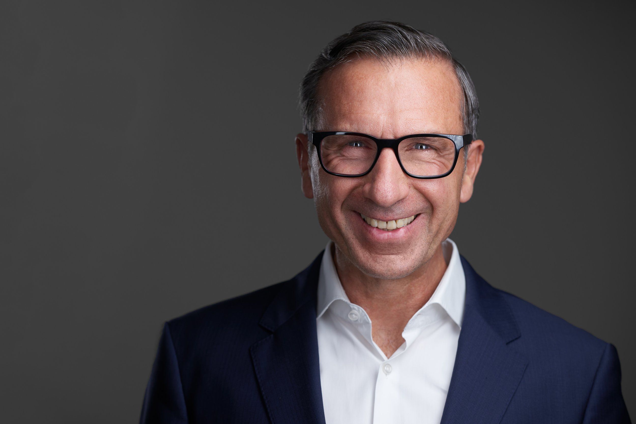 Businessportrait eines lächelnden Mannes mit Brille im dunkelblauen Anzug vor grauem Hintergrund, fotografiert im Studio in Köln