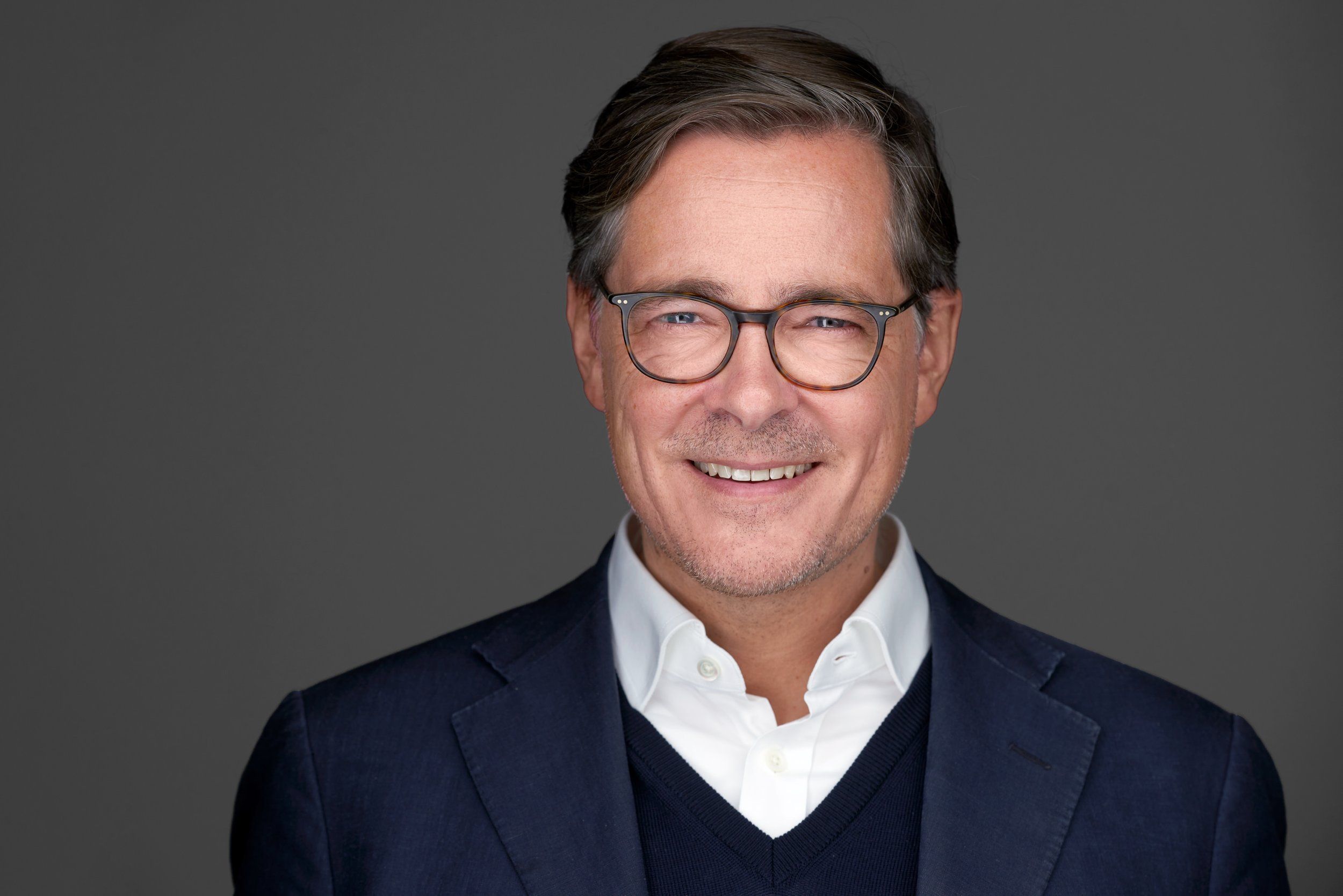 Businessportrait eines Mannes mit Brille im Sakko mit Pullover vor grauem Hintergrund, fotografiert im Studio in Köln
