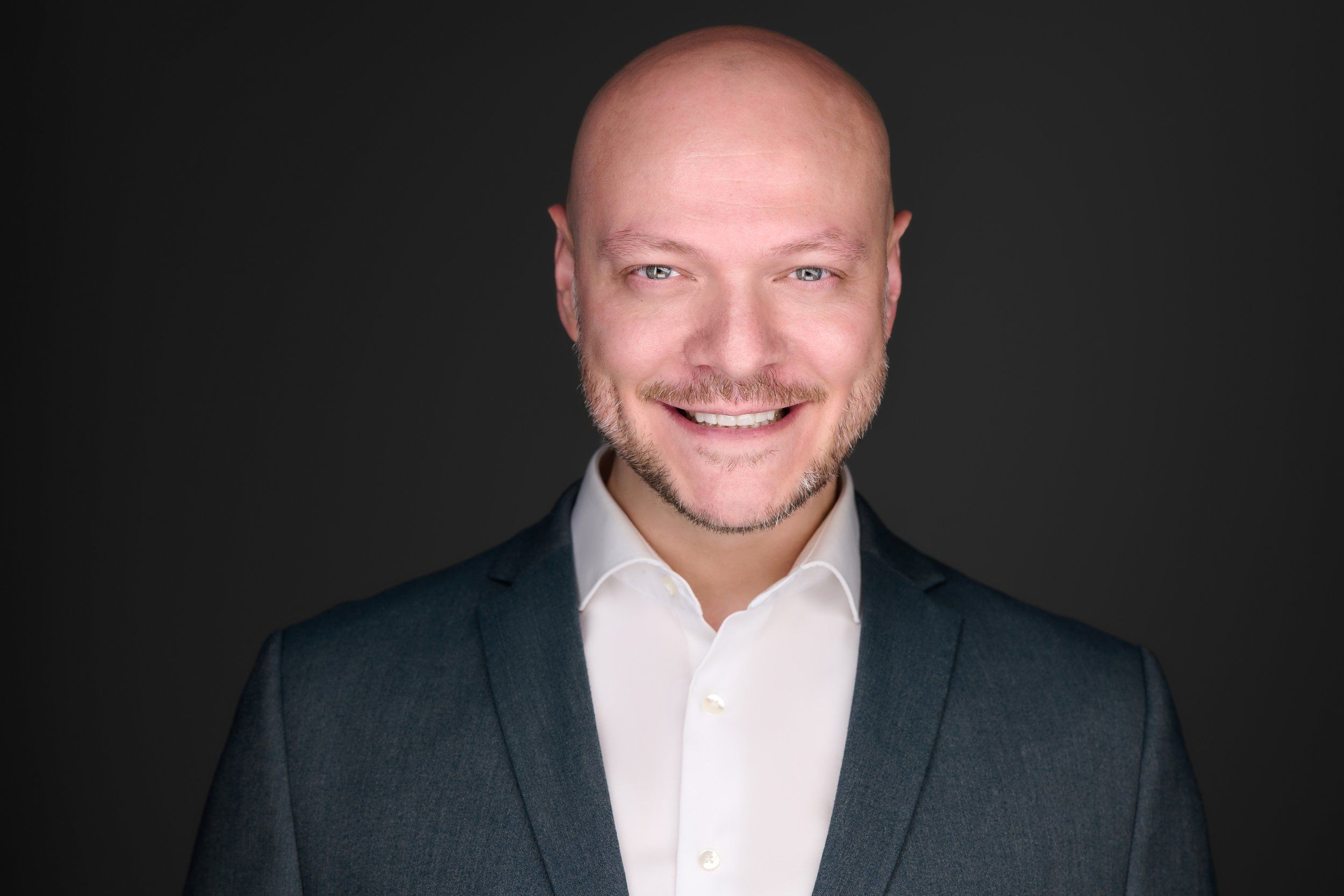 Businessportrait eines lächelnden Mannes mit Glatze im grauen Sakko vor dunklem Hintergrund, fotografiert im Studio in Köln