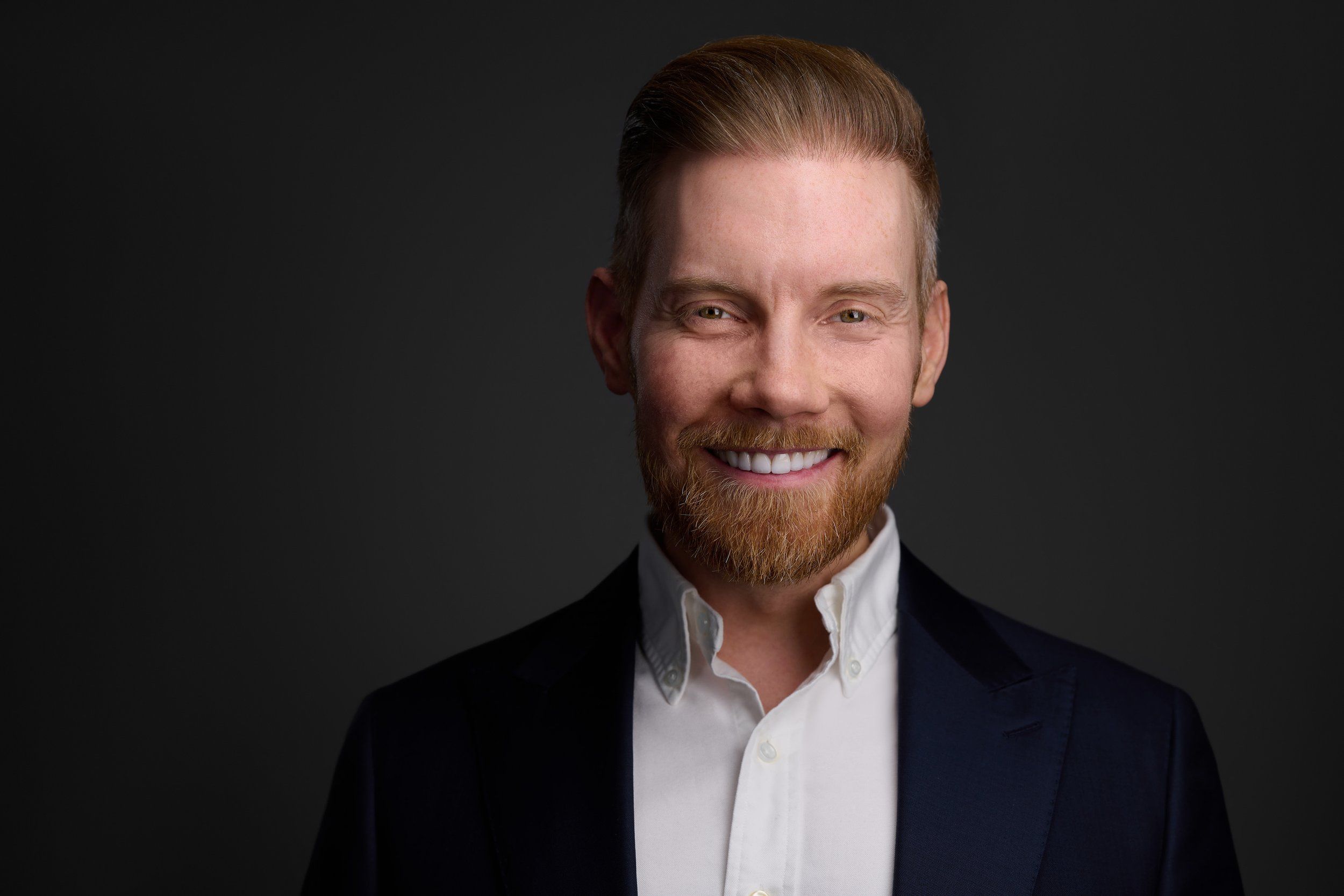 Businessportrait eines lächelnden Mannes mit rotblondem Bart im dunklen Sakko vor dunklem Hintergrund, fotografiert im Studio in Köln