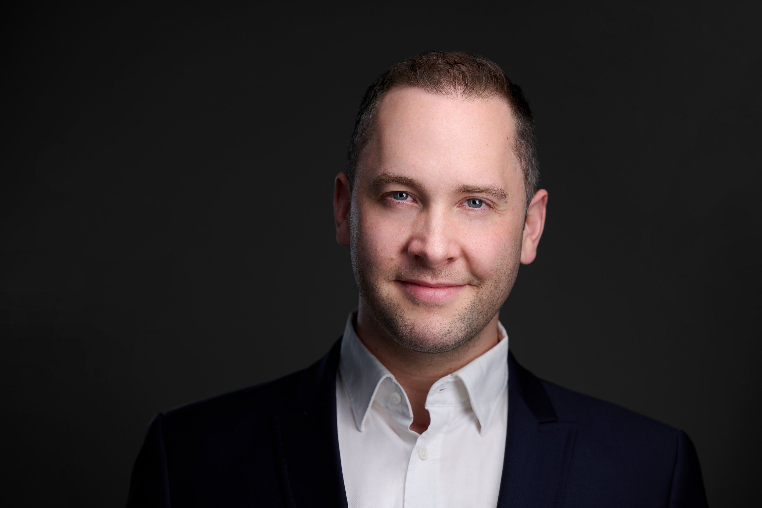 Businessportrait eines Mannes im schwarzen Sakko mit weißem Hemd vor dunklem Hintergrund, fotografiert im Studio in Köln