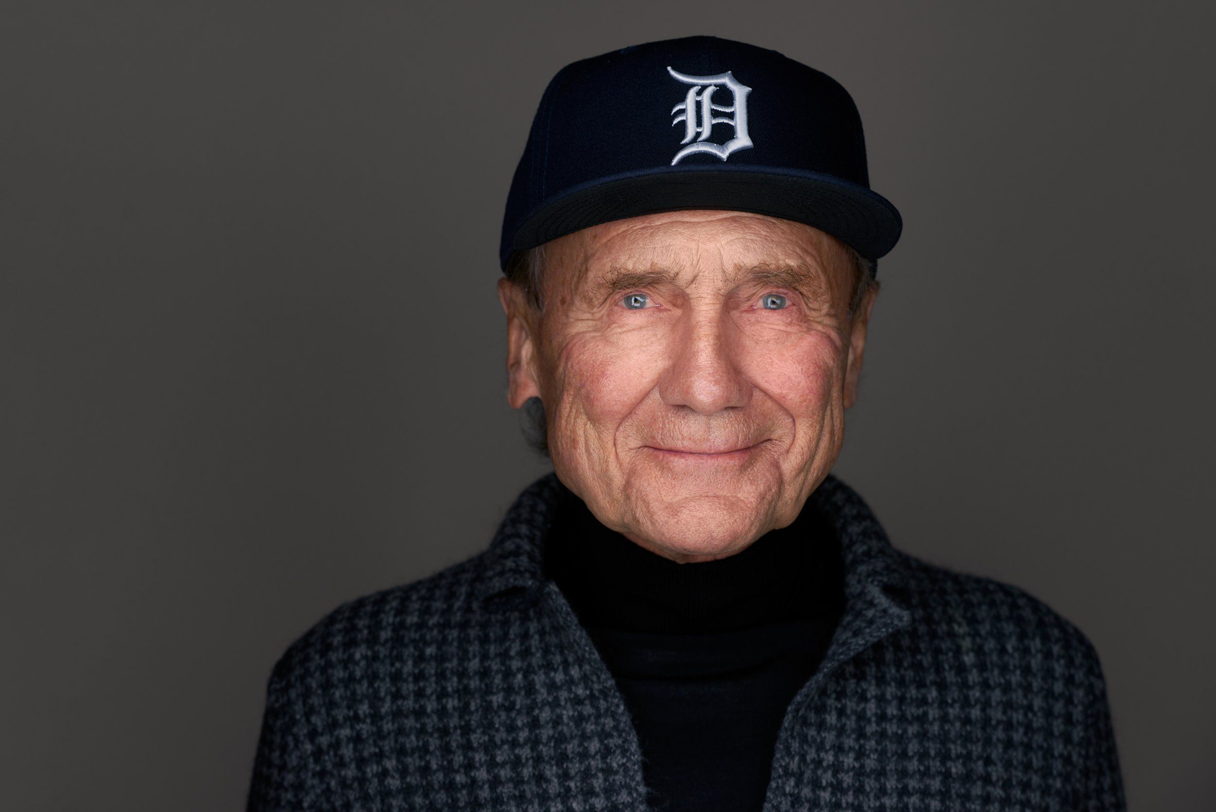 Headshot eines älteren, lächelnden Mannes mit Baseballcap und Rollkragen vor grauem Hintergrund, fotografiert im Studio in Köln