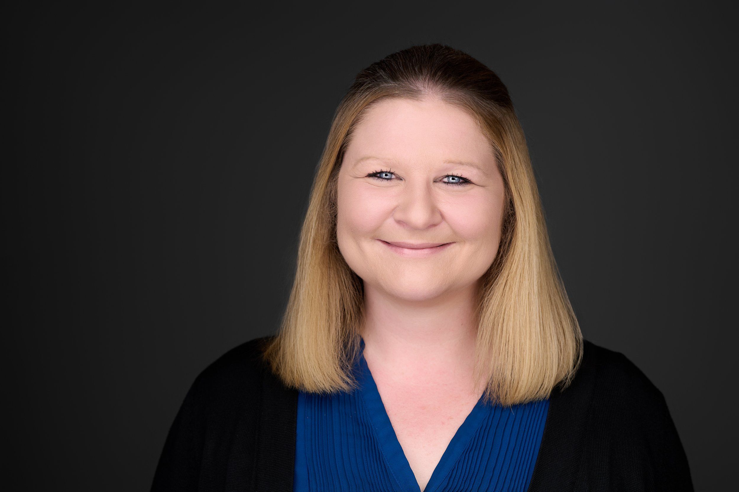 Headshot einer lächelnden Frau mit blonden Haaren in blauer Bluse vor dunklem Hintergrund, fotografiert im Studio in Köln