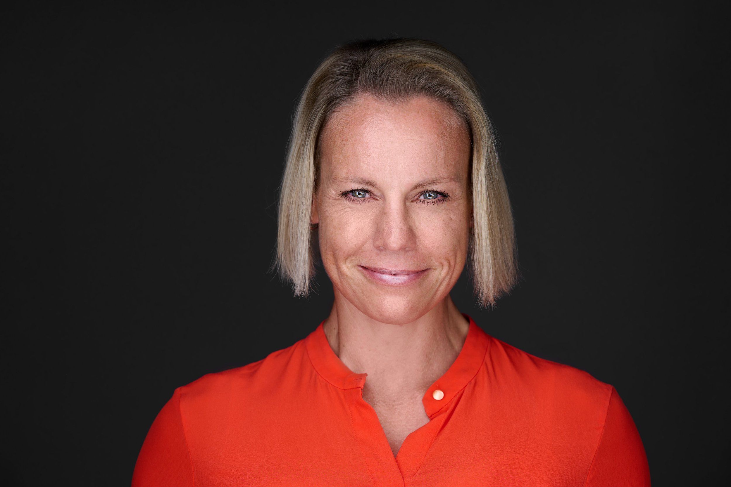 Headshot einer lächelnden Frau mit blondem Bob in orangefarbener Bluse vor dunklem Hintergrund, fotografiert im Studio in Köln