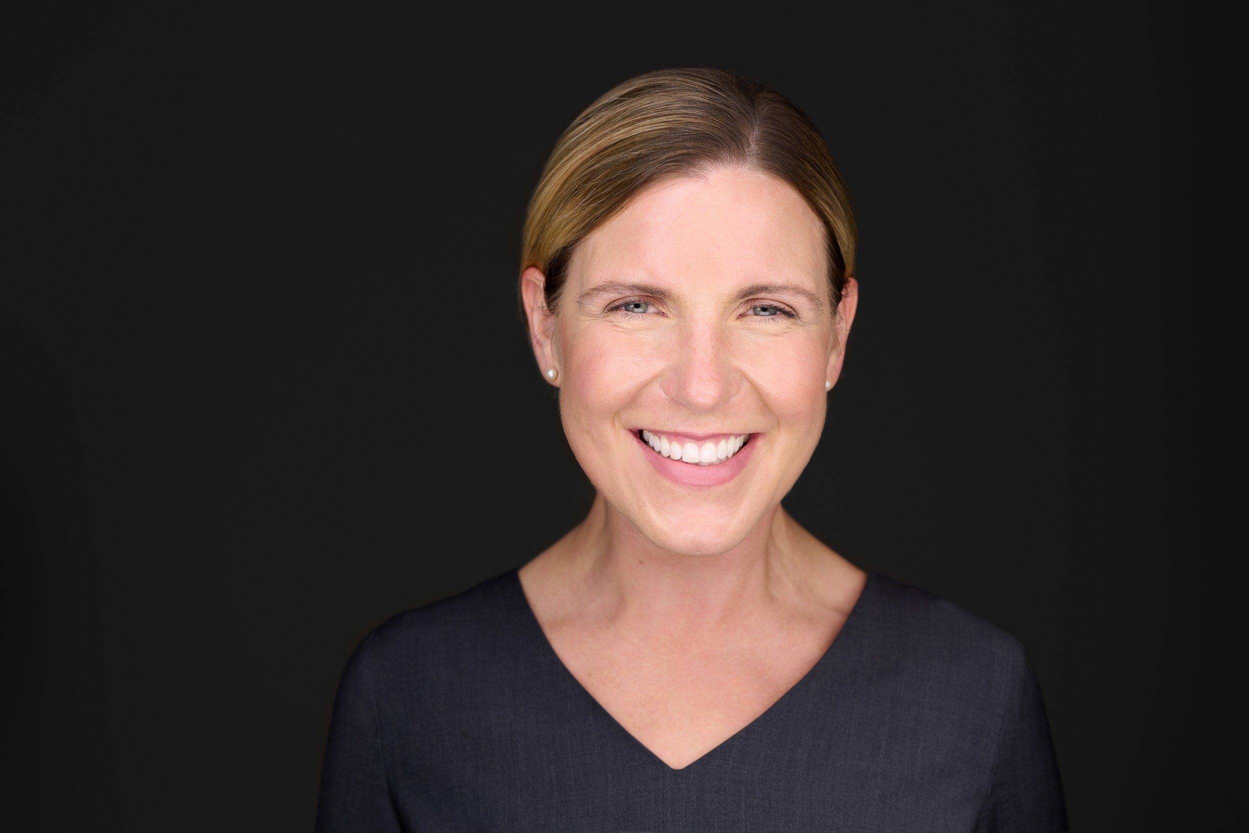 Headshot einer lächelnden Frau mit zurückgebundenen Haaren im grauen Oberteil vor dunklem Hintergrund, fotografiert im Studio in Köln