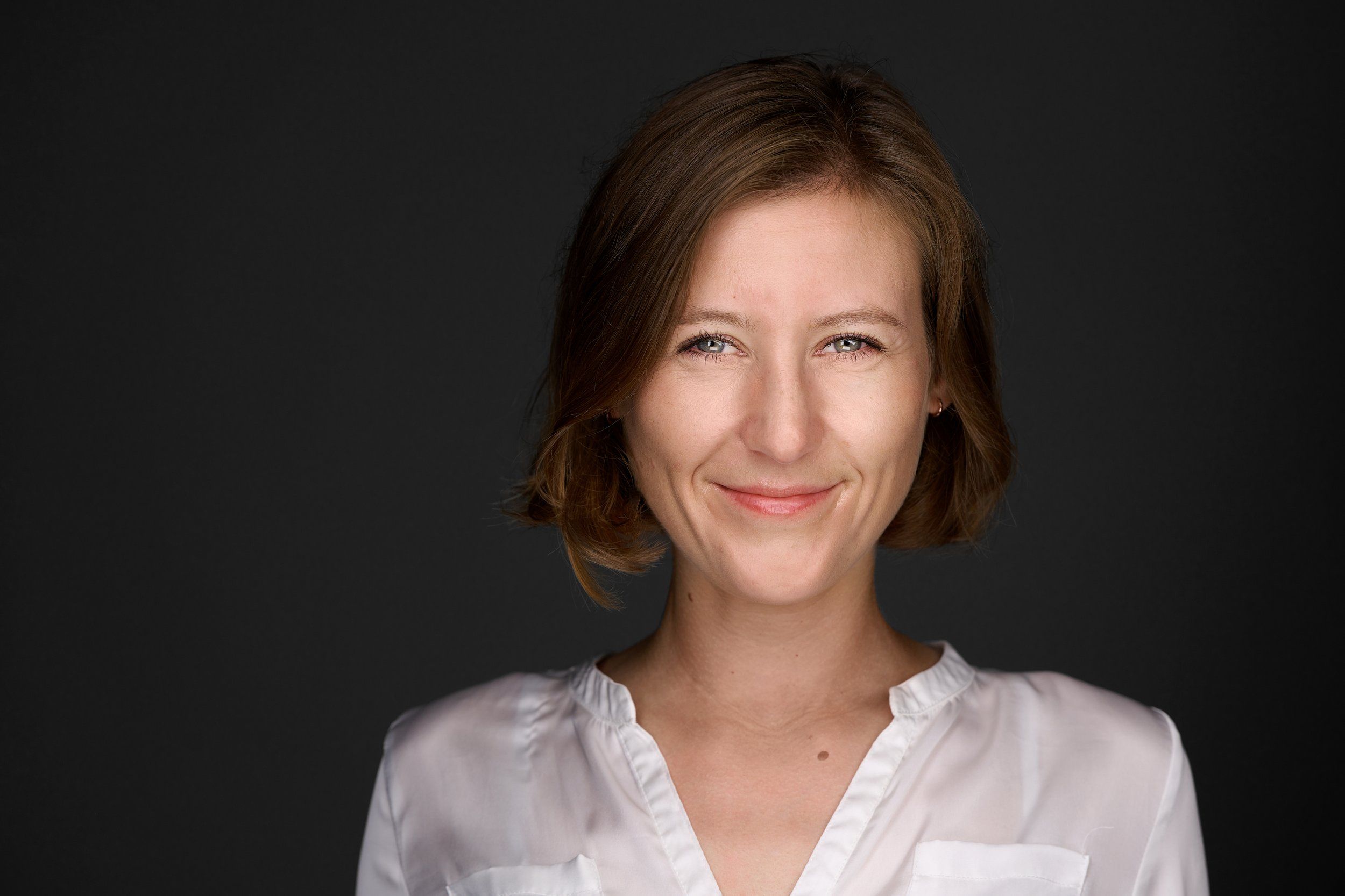 Headshot einer lächelnden Frau mit brünettem Bob in weißer Bluse vor dunklem Hintergrund, fotografiert im Studio in Köln