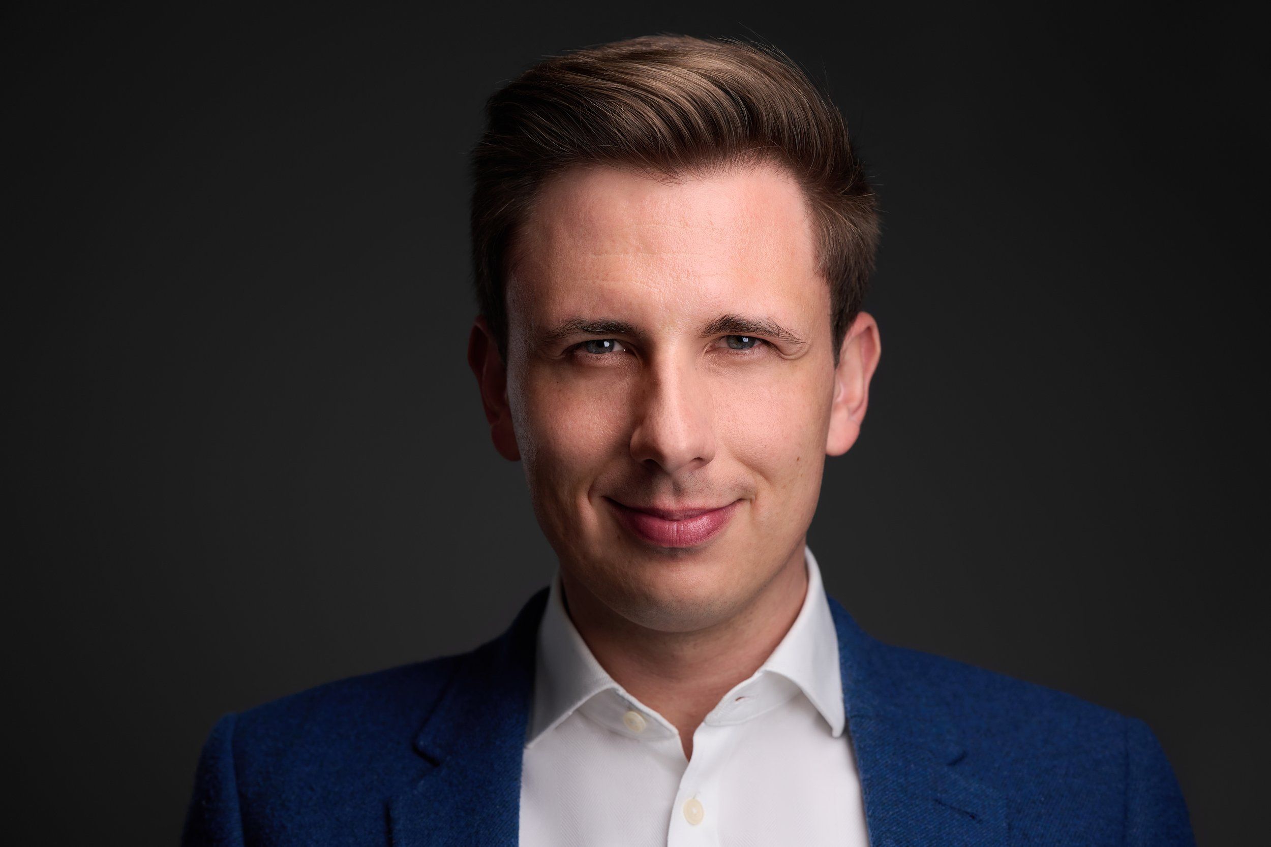 Headshot von Sebastian: junger Mann in blauem Sakko und weißem Hemd, lächelnd, vor dunklem Hintergrund
