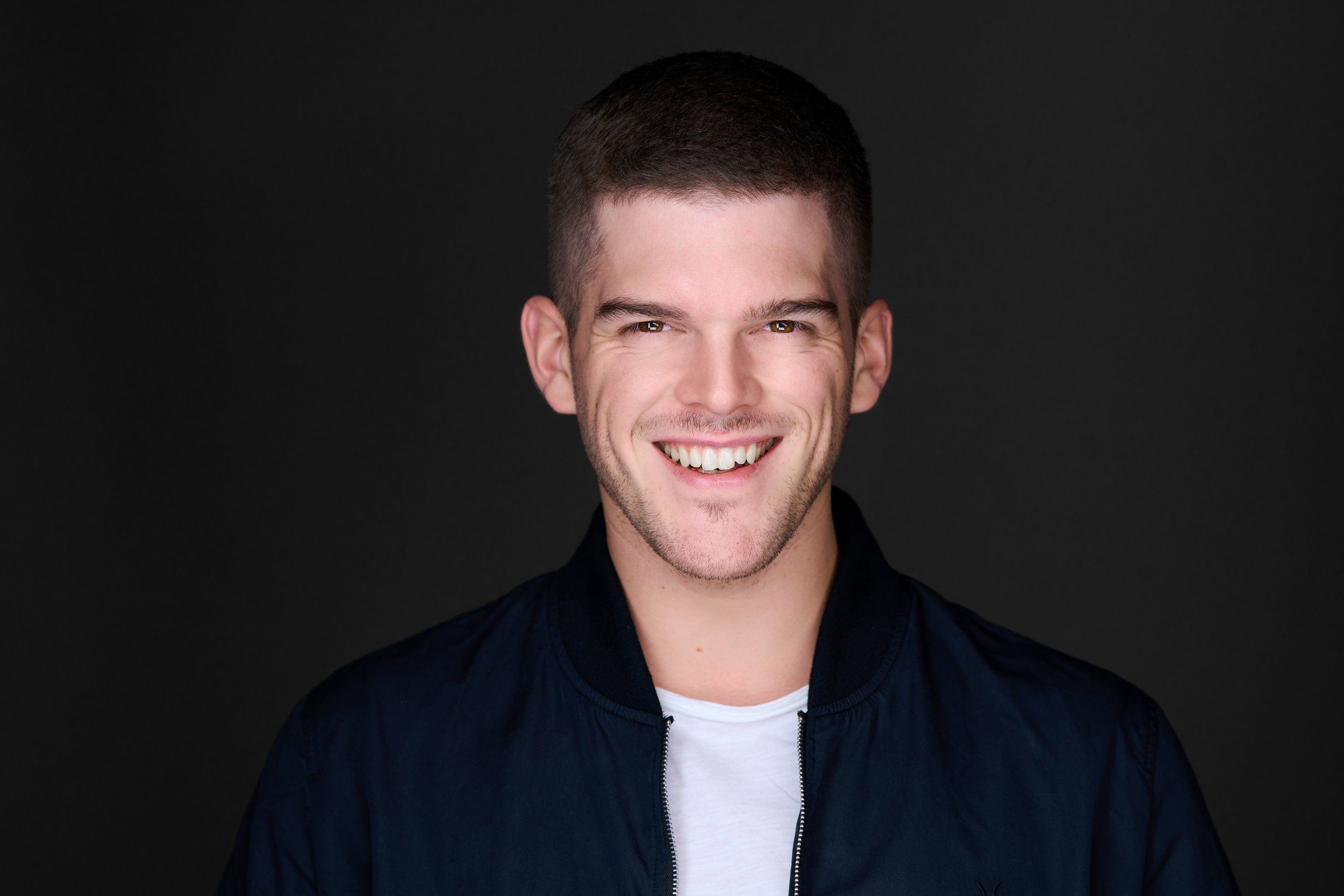 Headshot eines jungen, lachenden Mannes in dunkler Jacke und weißem T-Shirt vor dunklem Hintergrund, fotografiert im Studio in Köln
