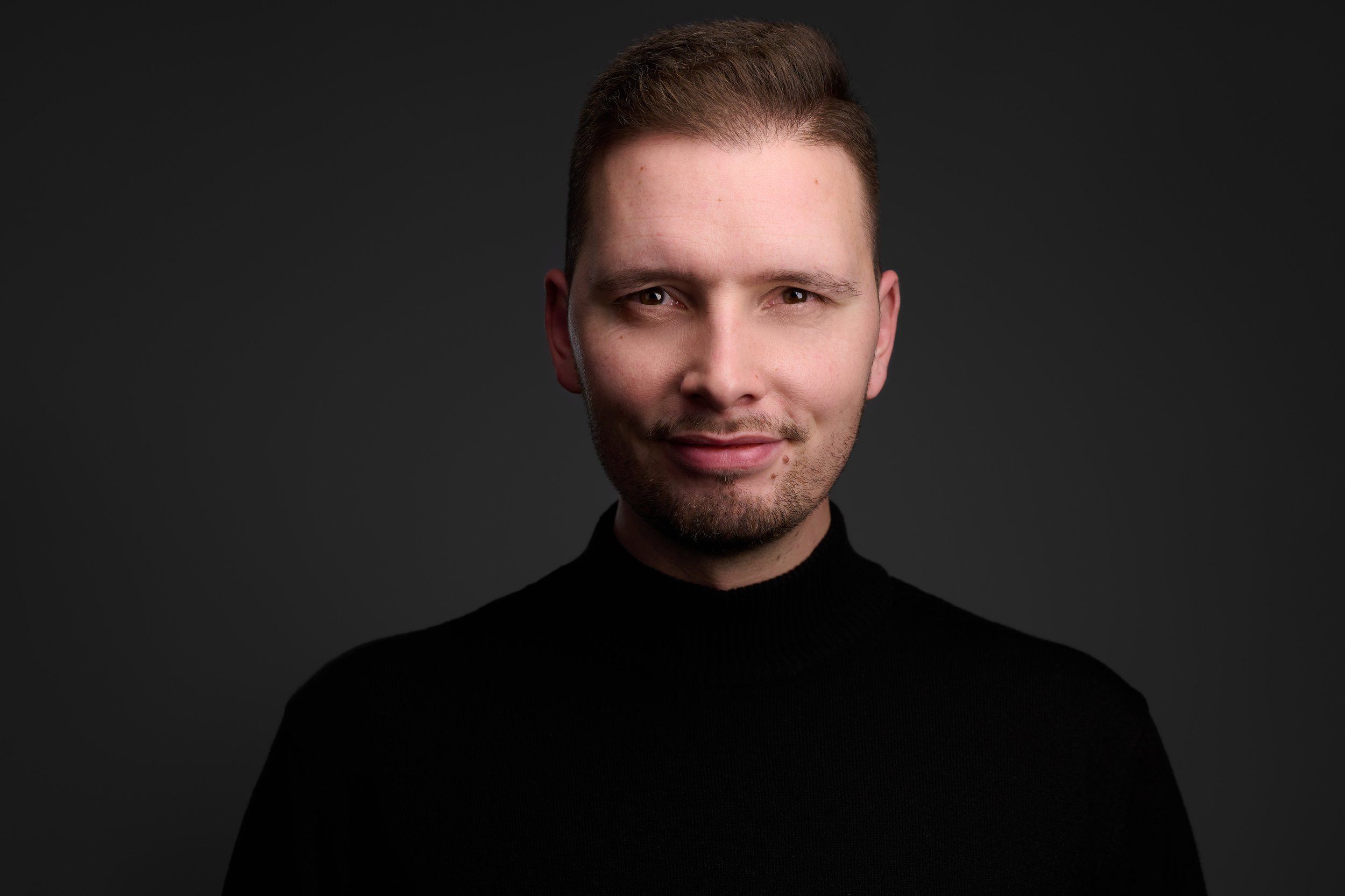 Headshot eines jungen Mannes im schwarzen Pullover vor schwarzem Hintergrund, fotografiert im Studio in Köln
