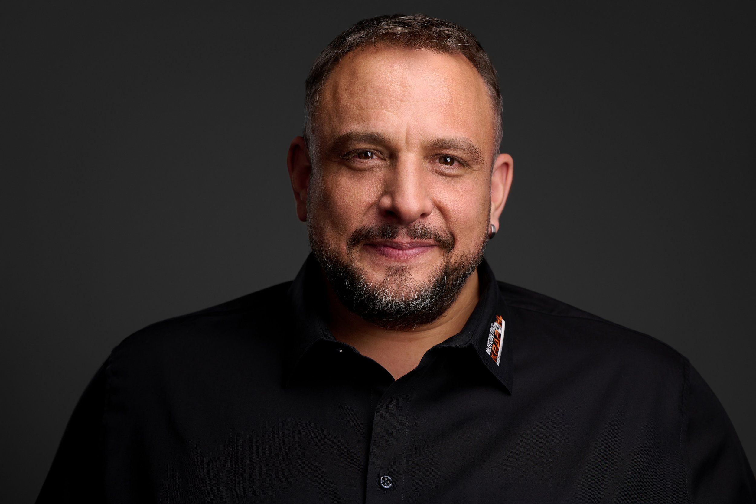 Headshot eines lächelnden Mannes mit Bart im schwarzen Hemd vor dunklem Hintergrund, fotografiert im Studio in Köln