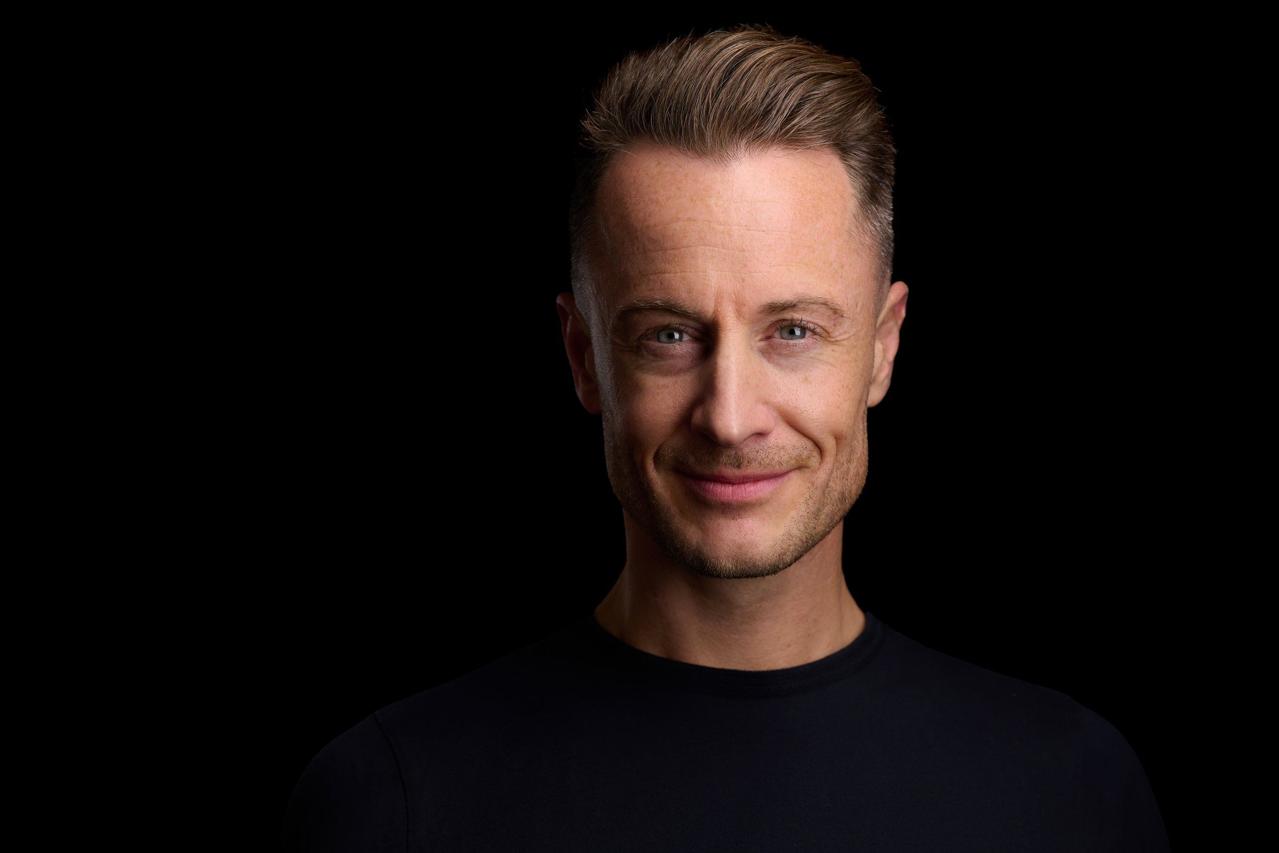 Headshot eines blonden Mannes im schwarzen Pullover vor schwarzem Hintergrund, fotografiert im Studio in Köln