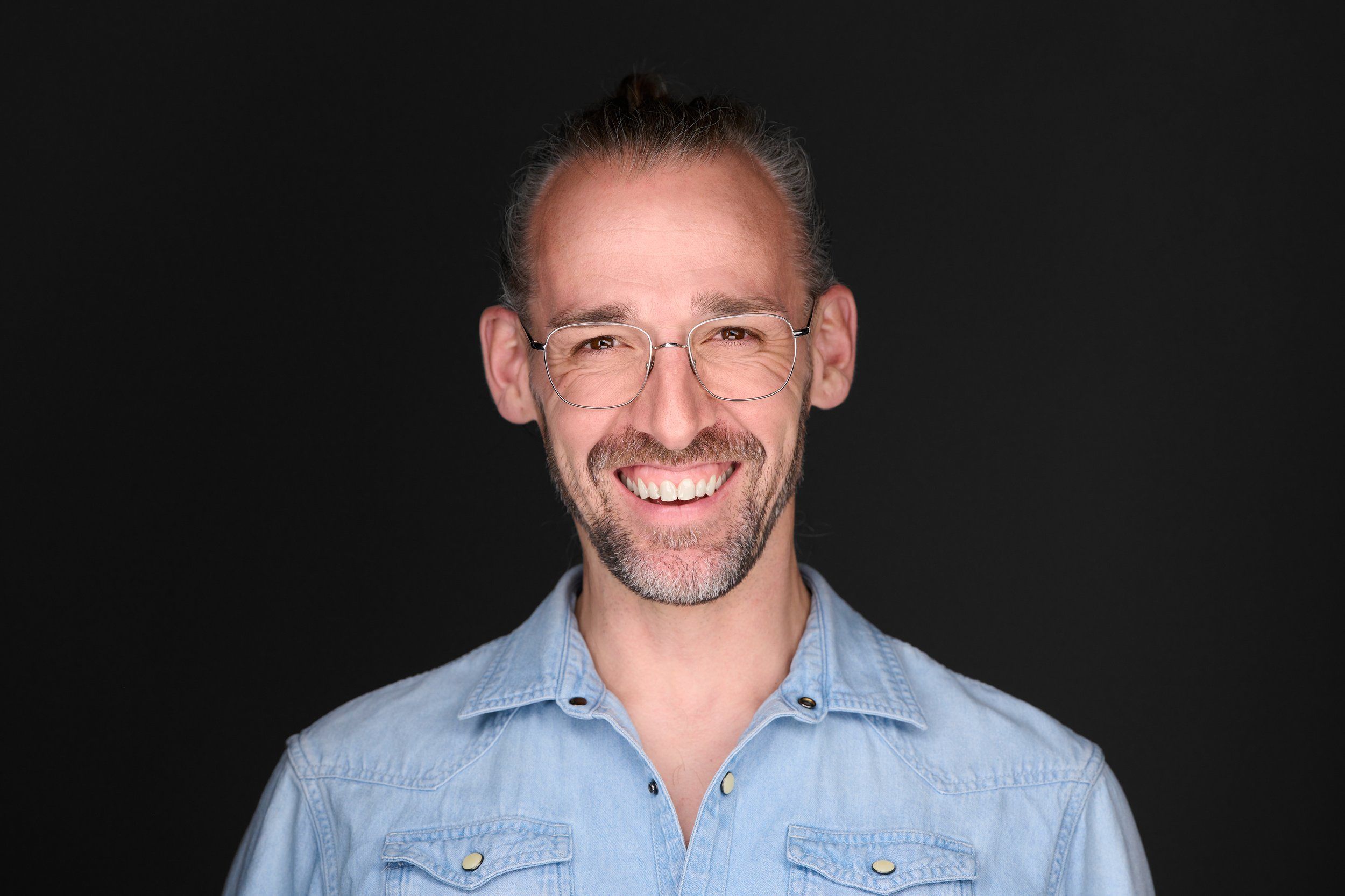 Headshot eines lachenden Mannes mit Brille im Jeanshemd vor dunklem Hintergrund, fotografiert im Studio in Köln