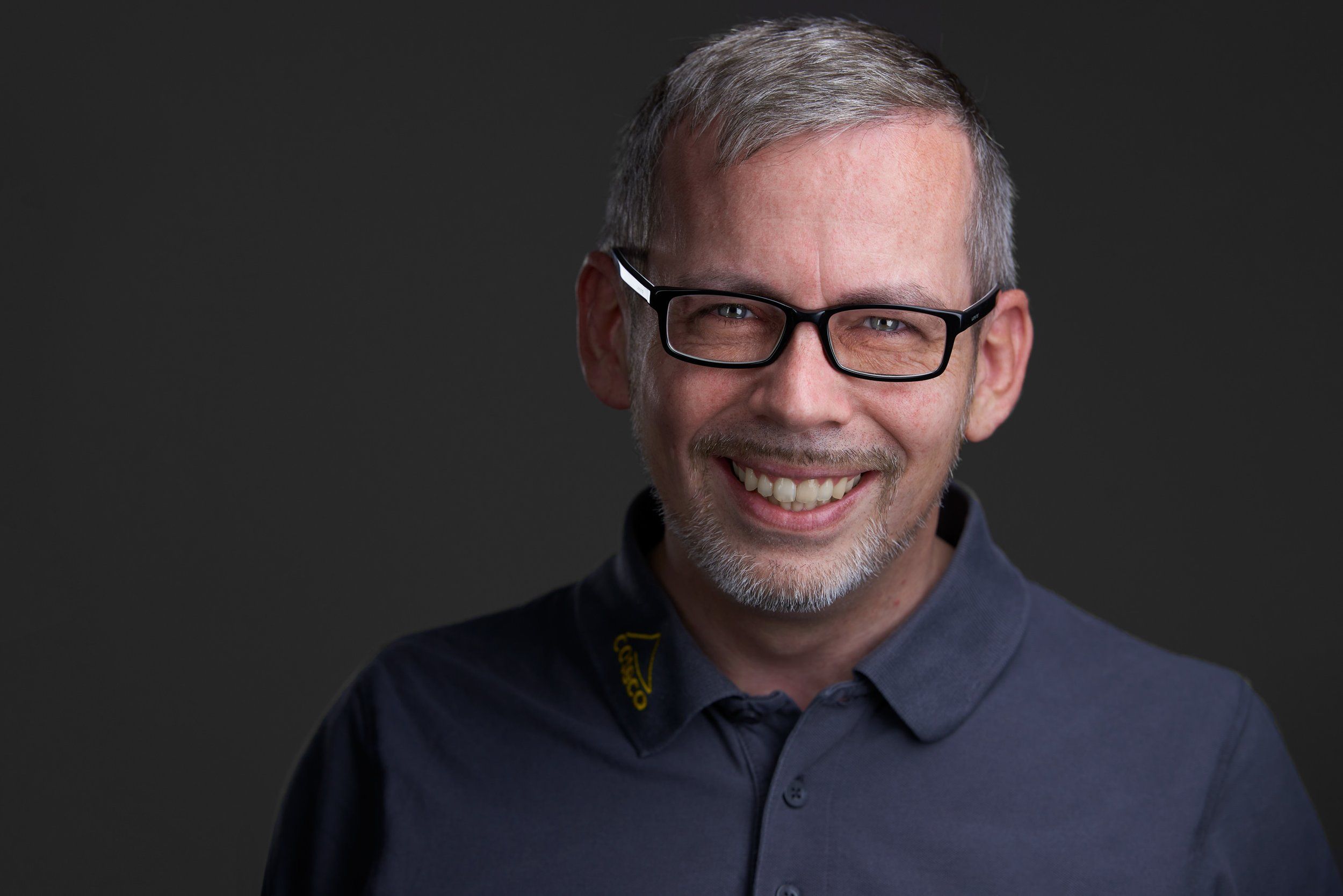 Headshot eines lachenden Mannes mit Brille und grauen Haaren im Poloshirt vor dunklem Hintergrund, fotografiert im Studio in Köln