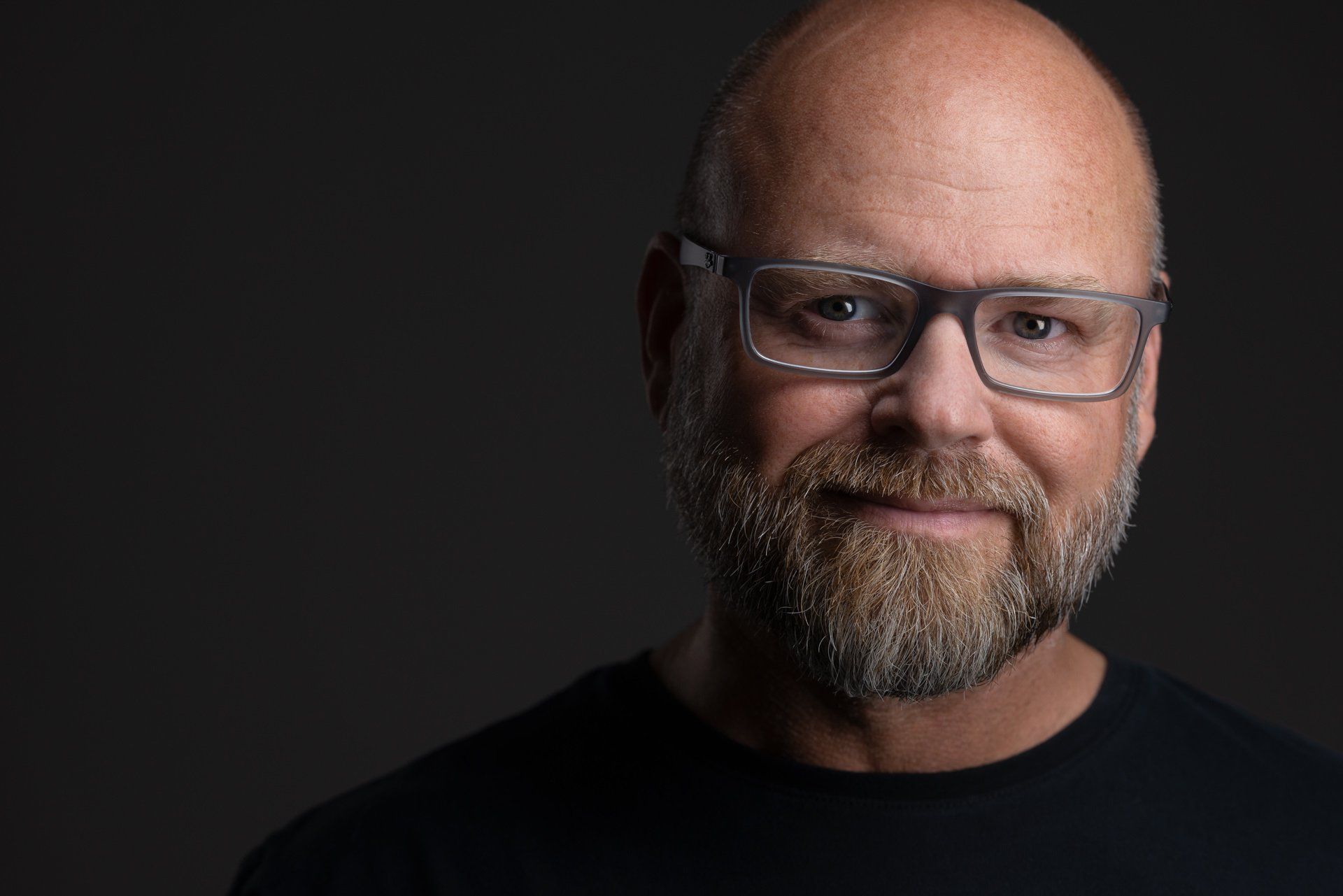 Headshot eines Mannes mit Glatze, Brille und Vollbart im schwarzen Shirt vor dunklem Hintergrund, fotografiert im Studio in Köln