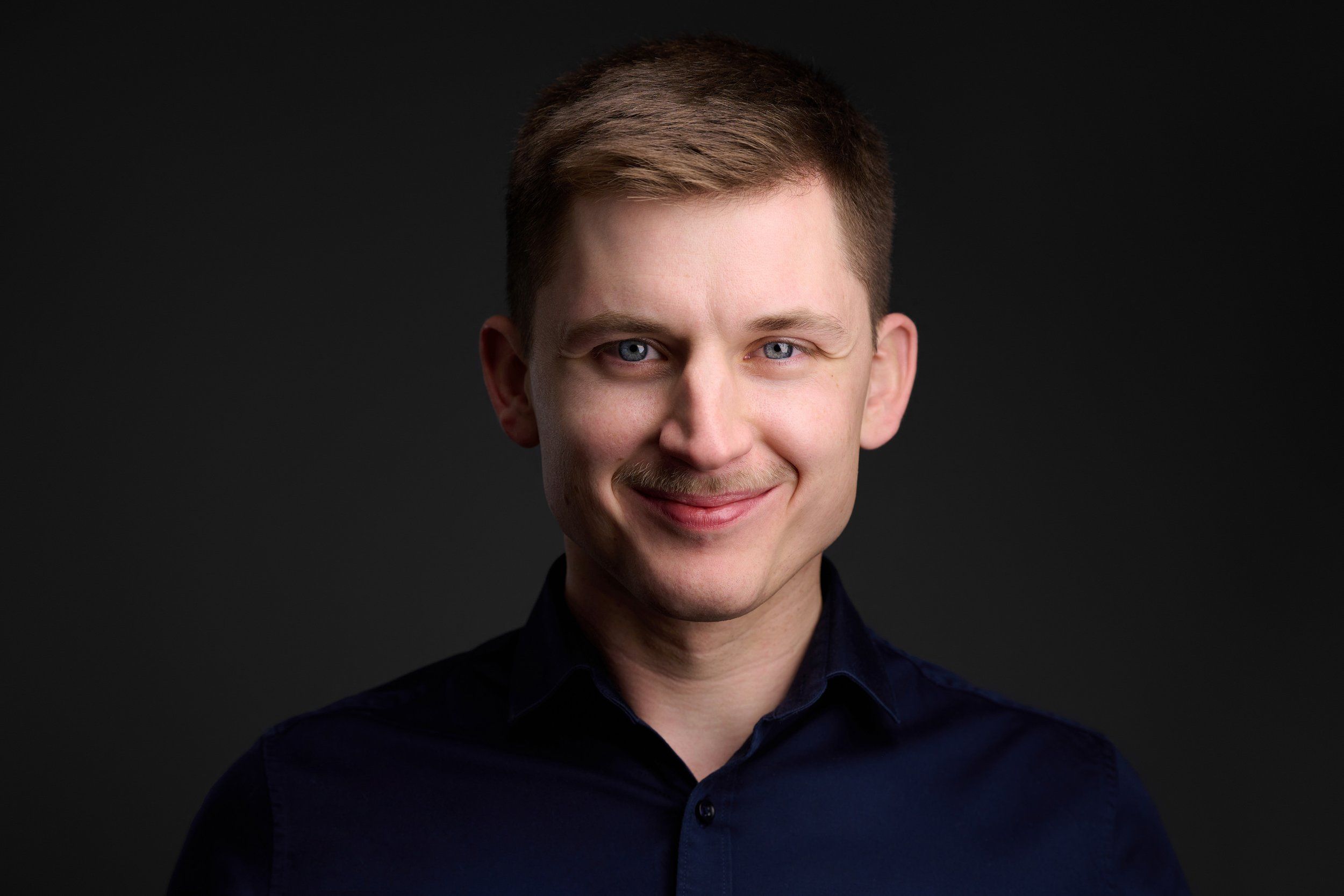 Headshot eines lächelnden Mannes im dunkelblauen Hemd vor dunklem Hintergrund, fotografiert im Studio in Köln
