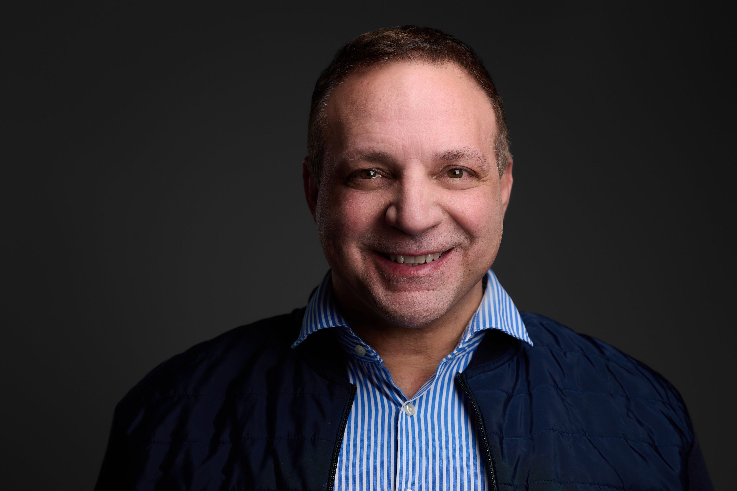 Headshot eines lächelnden Mannes mit gestreiftem Hemd und dunkler Steppjacke vor dunklem Hintergrund, fotografiert im Studio in Köln