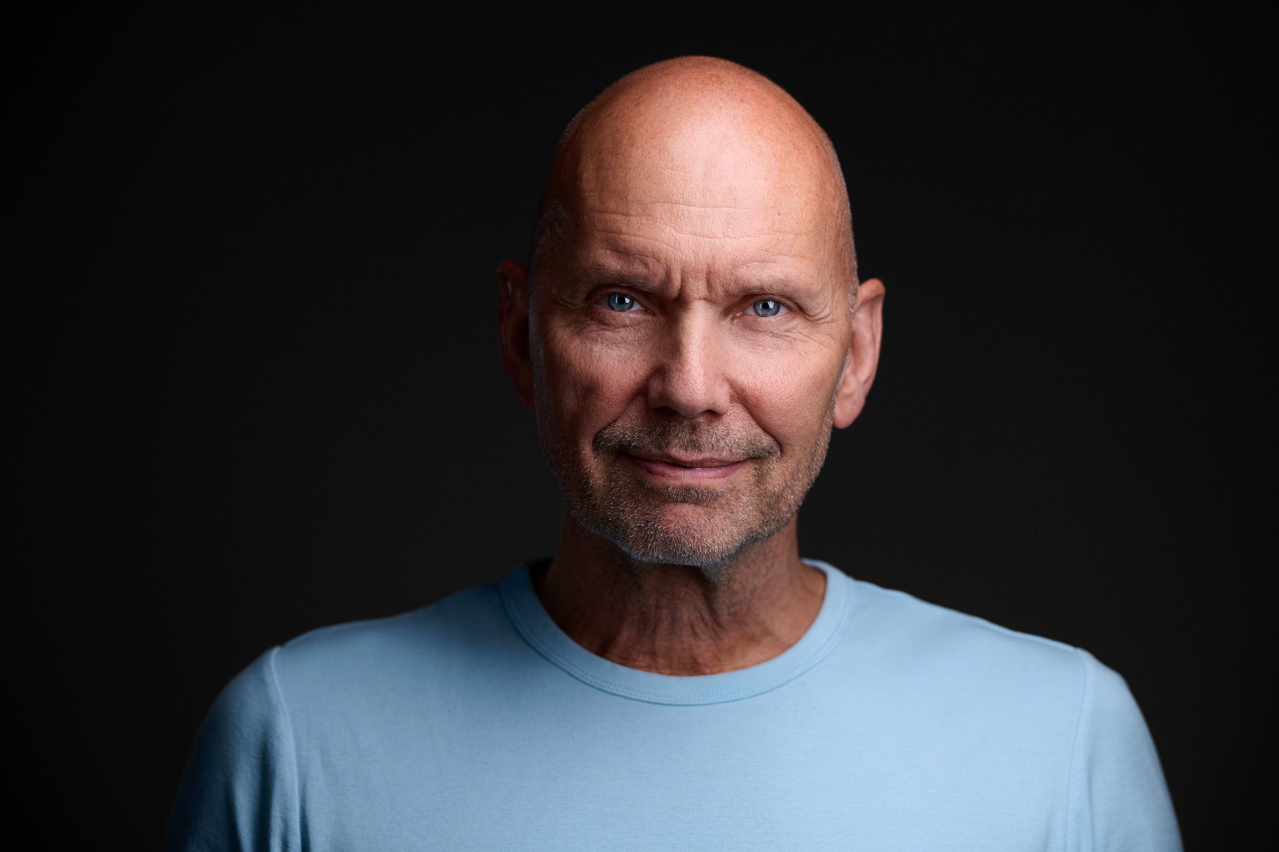Headshot eines Mannes mit Glatze im hellblauen T-Shirt vor schwarzem Hintergrund, fotografiert im Studio in Köln