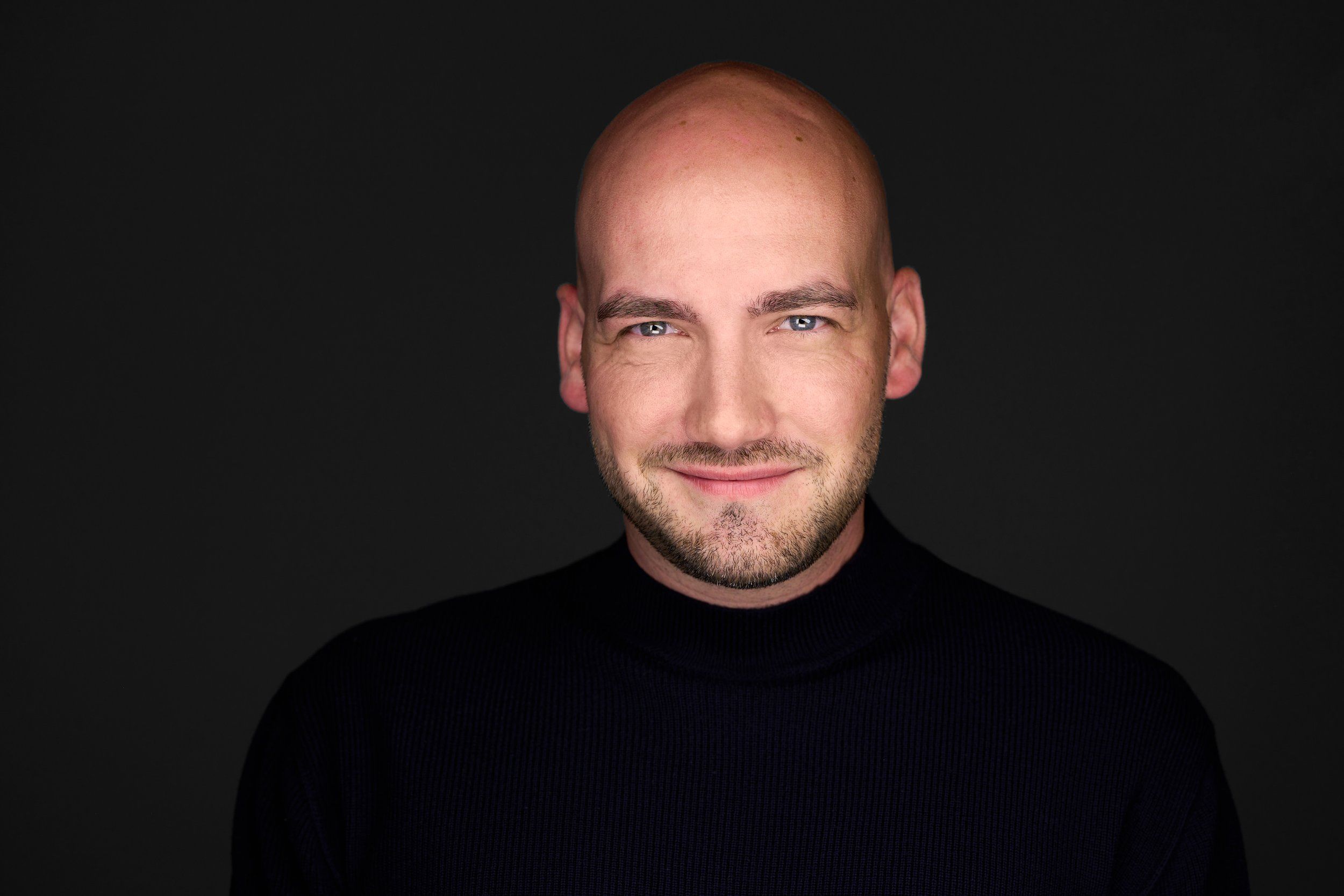 Headshot eines Mannes mit Glatze und Bart im schwarzen Rollkragenpullover vor schwarzem Hintergrund, fotografiert im Studio in Köln