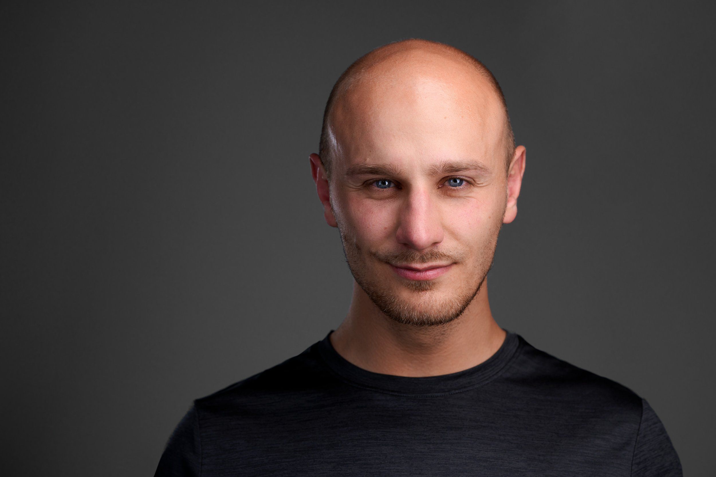 Headshot eines Mannes mit Glatze im schwarzen Shirt vor dunkelgrauem Hintergrund, fotografiert im Studio in Köln