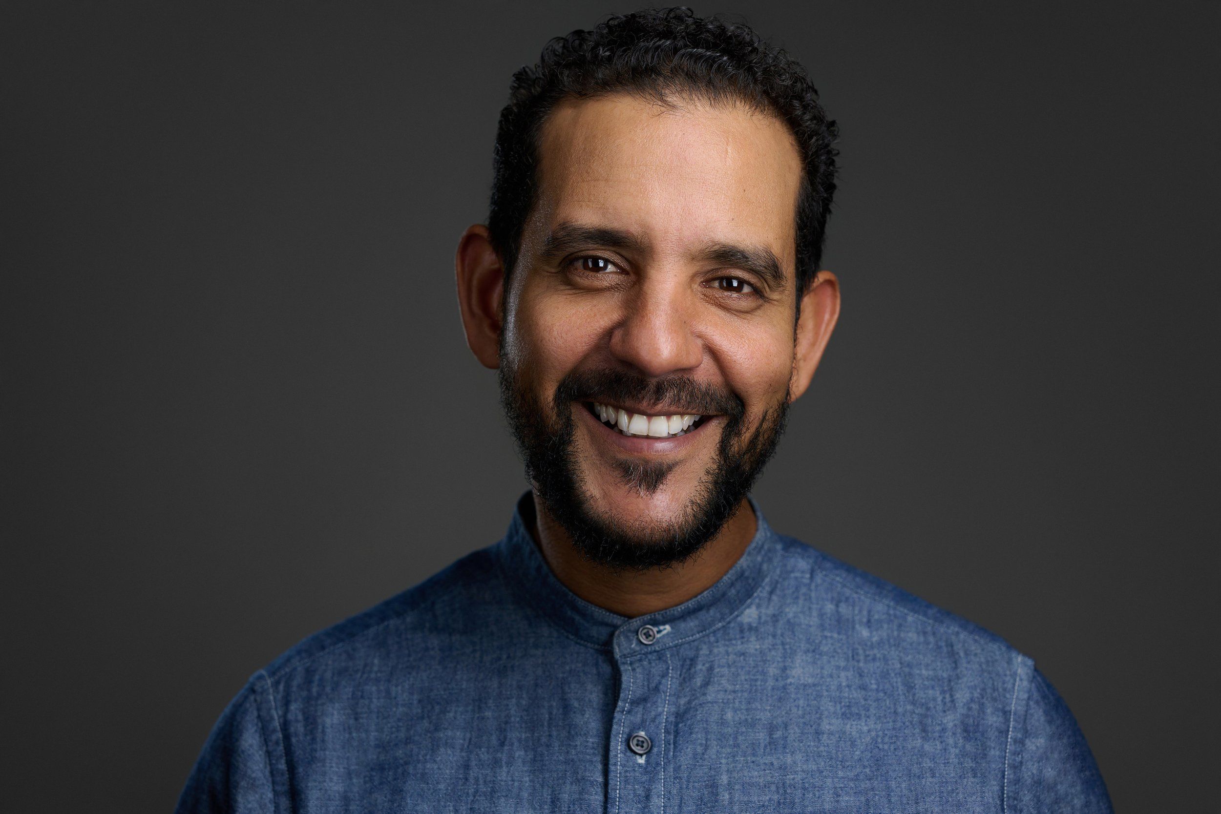 Headshot eines lachenden Mannes mit Bart im blauen Hemd vor dunklem Hintergrund, fotografiert im Studio in Köln