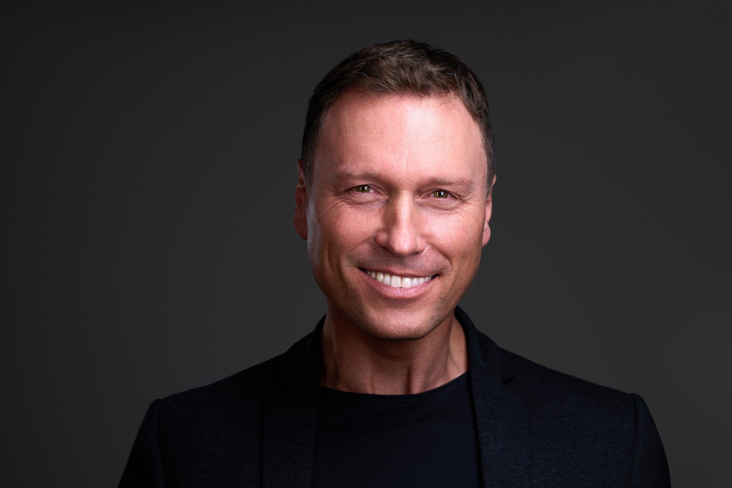 Headshot eines lächelnden Mannes im schwarzen Sakko vor dunkelgrauem Hintergrund, fotografiert im Studio in Köln