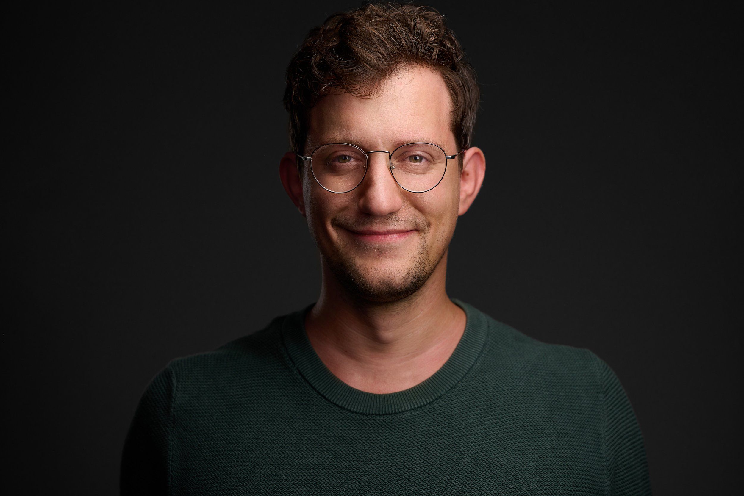 Headshot eines Mannes mit runder Brille im grünen Pullover vor dunklem Hintergrund, fotografiert im Studio in Köln