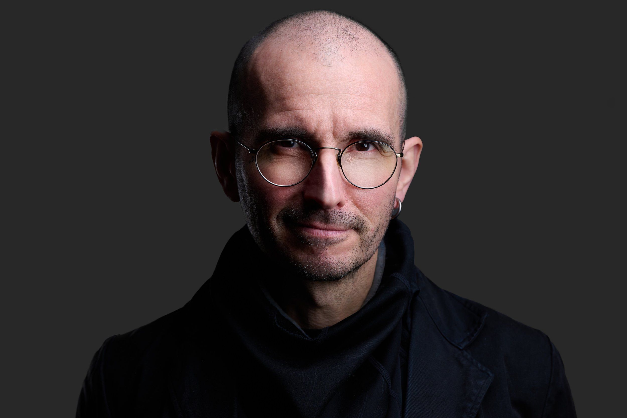 Headshot eines Mannes mit Glatze und runder Brille in schwarzer Kleidung vor dunklem Hintergrund, fotografiert im Studio in Köln