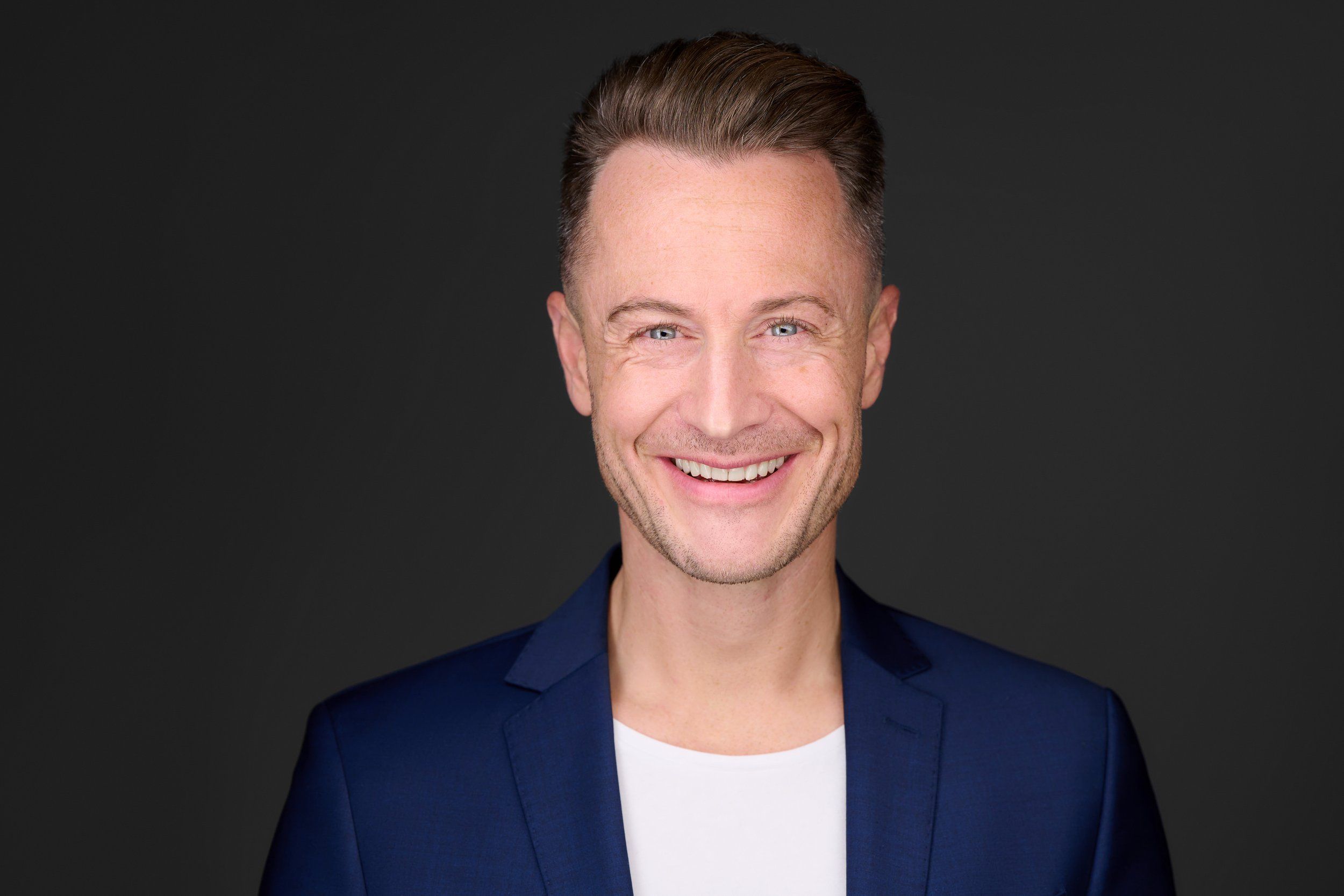 Headshot von Stefan: lachender Mann in blauem Sakko und weißem T-Shirt vor schwarzem Hintergrund