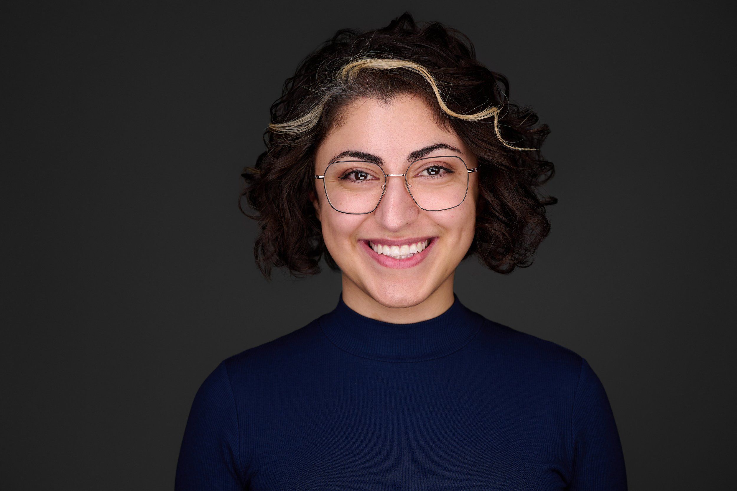 Headshot einer lächelnden Frau mit Locken und Brille im dunkelblauen Pullover vor dunklem Hintergrund, fotografiert im Studio in Köln