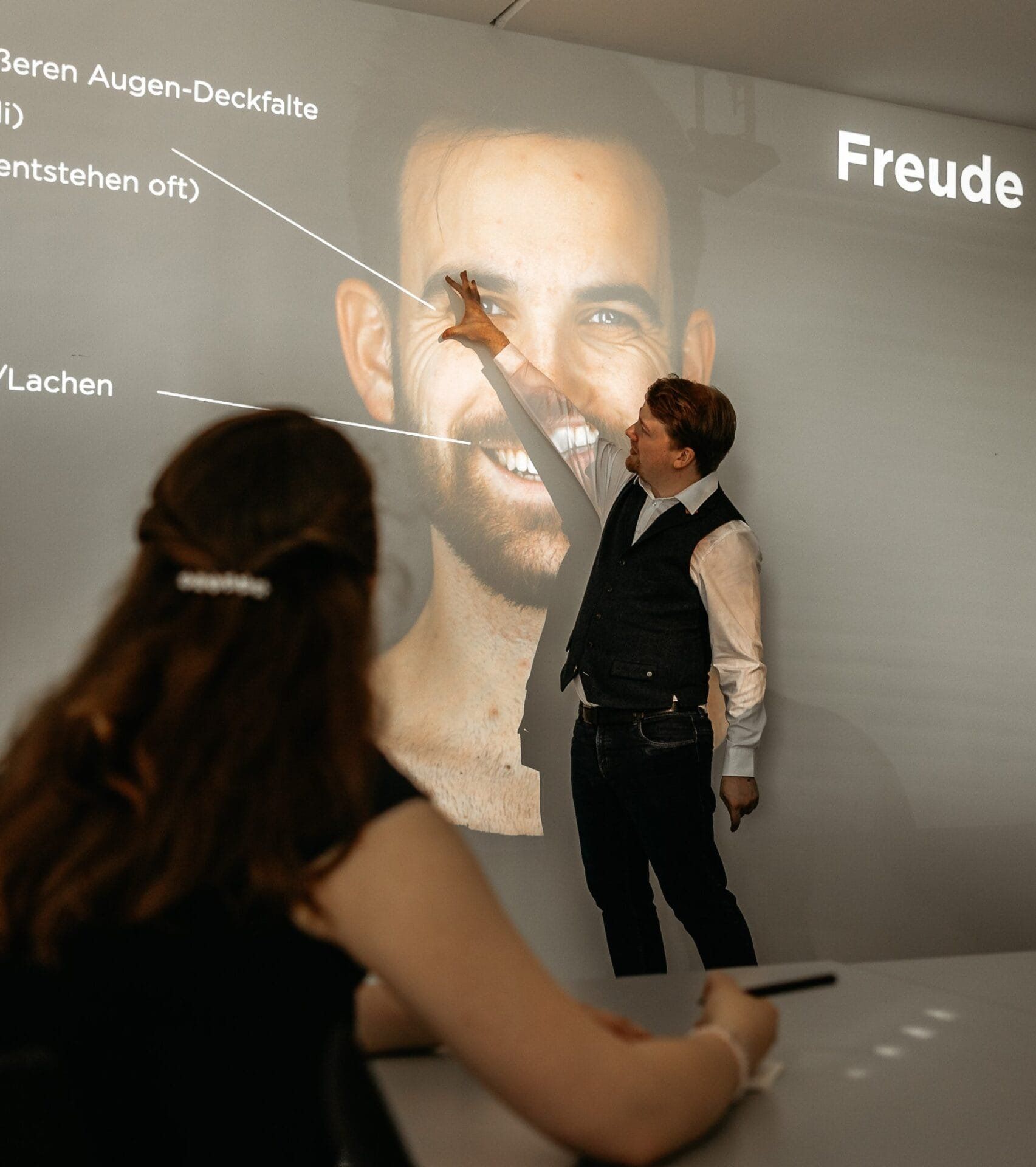 Oliver Wagner erklärt im Seminar Gesichtsausdrücke an einer Beamer-Projektion, eine Teilnehmerin macht sich Notizen